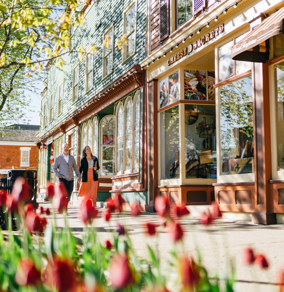 An older couple walking down the side walk looking into the windows of boutique shops in Niagara-on-the-Lake. Photo is taken peeking through a bed of tulips.